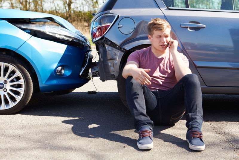 A young man sits next to two crashed cars and talks on the phone.