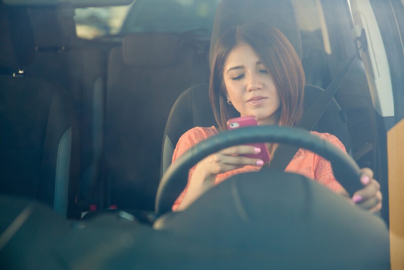 A young woman wearing a black blazer is looking at her phone while sitting in the driver's seat of a car.