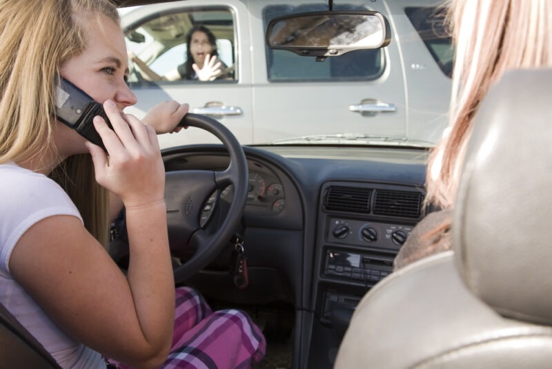 A driver has one hand on the steering wheel while holding her phone to her ear with the other hand.