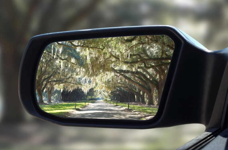 A side rear view mirror on a car showing a Charleston-area country road with trees around it.