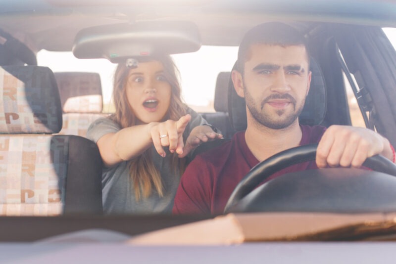 A man is driving a car while a woman in the back seat looks surprised and is pointing.