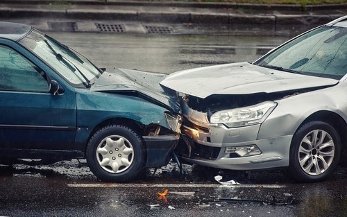 Two cars damaged in a collision, with visible dents and broken parts, parked on the side of the road.